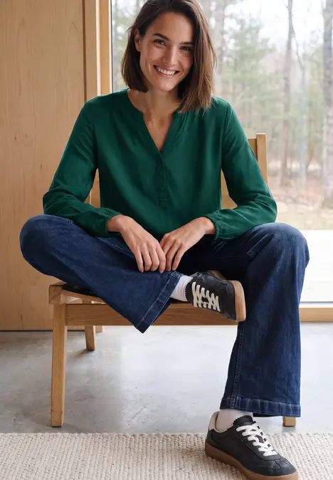 Eine lächelnde Frau sitzt im Schneidersitz auf einem Stuhl, trägt grüne Bluse, Jeans und schwarze Sneaker.