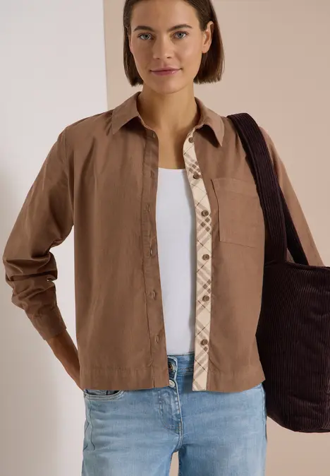 Eine Frau mit braunem Hemd, weißem Top, Jeans und schwarzer Tasche vor hellem Hintergrund.