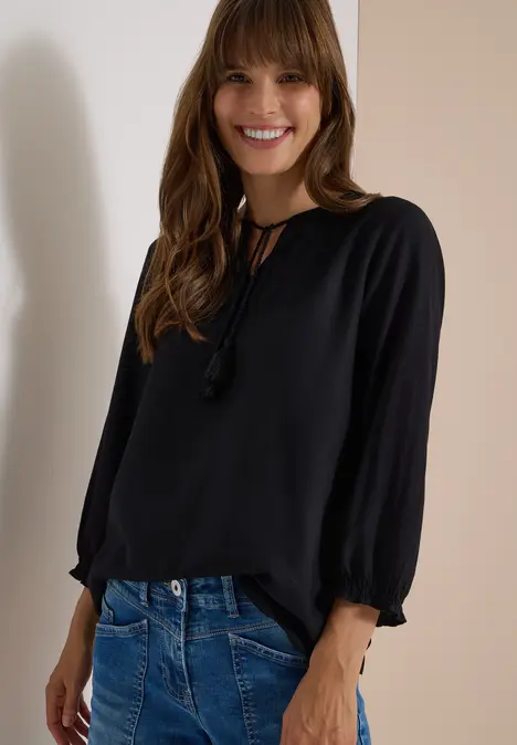 Eine lächelnde Frau trägt eine schwarze Bluse mit Quasten am Ausschnitt und lockere blaue Jeans.