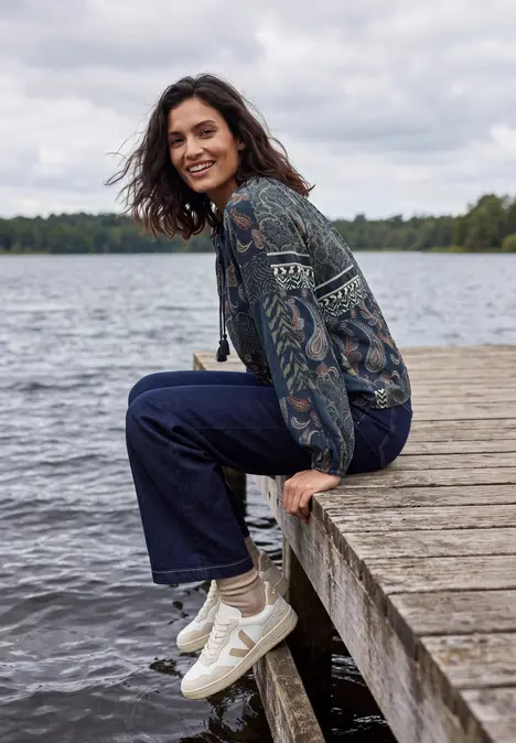Eine Frau sitzt auf einem Steg am See, trägt Jeans, gemusterte Jacke und weiße Sneaker, lächelt entspannt.