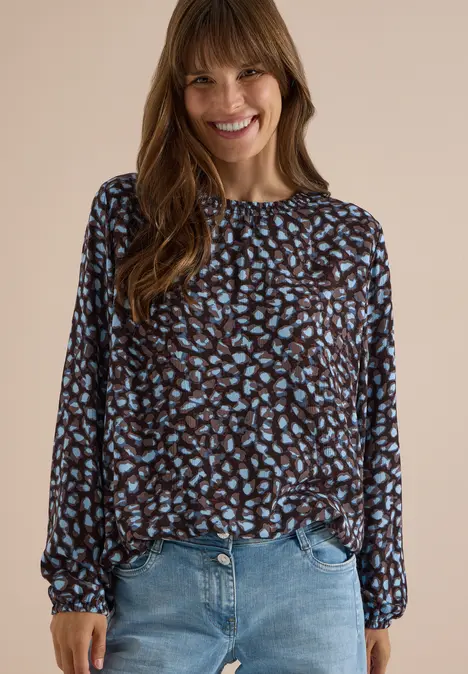 Eine lächelnde Frau trägt eine braun-schwarze Bluse mit blauem Blattmuster und Jeans vor beigem Hintergrund.
