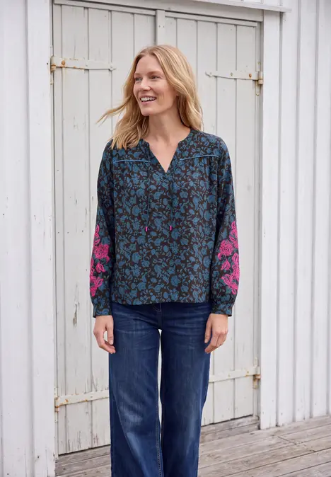 Eine lächelnde Frau in blau gemusterter Bluse mit pinken Ärmeln und Jeans vor einer weißen Holzwand.