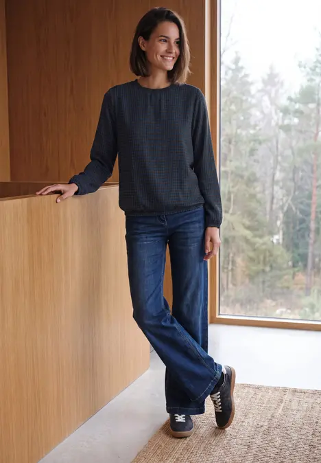 Eine lächelnde Frau in Jeans und dunklem Pullover lehnt an einer Holztheke und schaut aus dem Fenster.