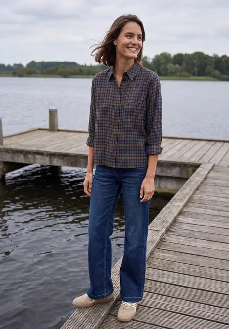 Frau in kariertem Hemd und Jeans steht lächelnd auf einem Holzsteg am See.