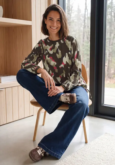 Eine Frau sitzt lächelnd auf einem Stuhl in einem hellen Raum, trägt Camouflage-Pullover und Jeans.