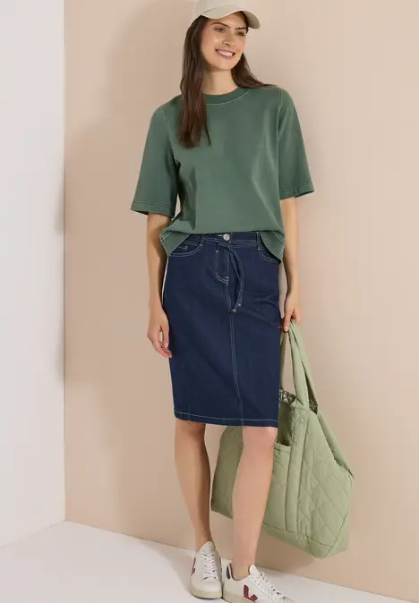Eine Frau trägt ein grünes T-Shirt, einen dunkelblauen Denim-Rock, weiße Sneaker und hält eine gesteppte grüne Tasche.