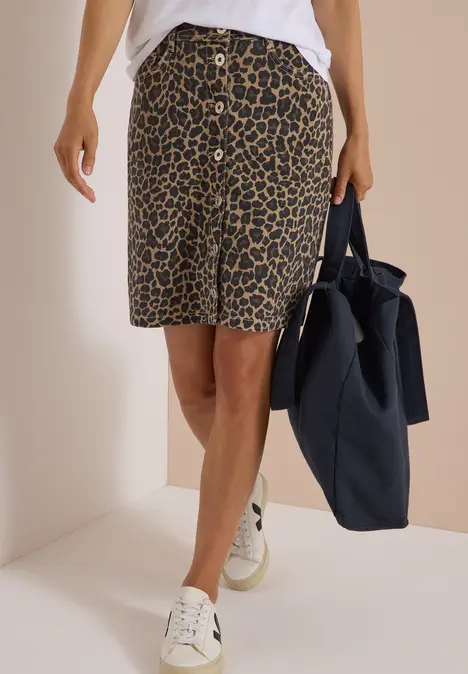 Iemand draagt een knielange rok met luipaardprint, witte sneakers en houdt een donkerblauwe tas vast.