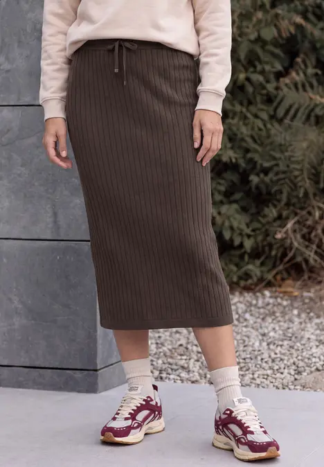 Person trägt hellen Pullover, braunen, gerippten Midirock und weiße Sneaker mit roten Details vor Betonwand.