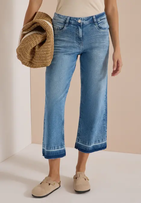 Person trägt weite, knöchellange Jeans mit Umschlag, beiges Top, Schlupfschuhe und hält Strohhut.