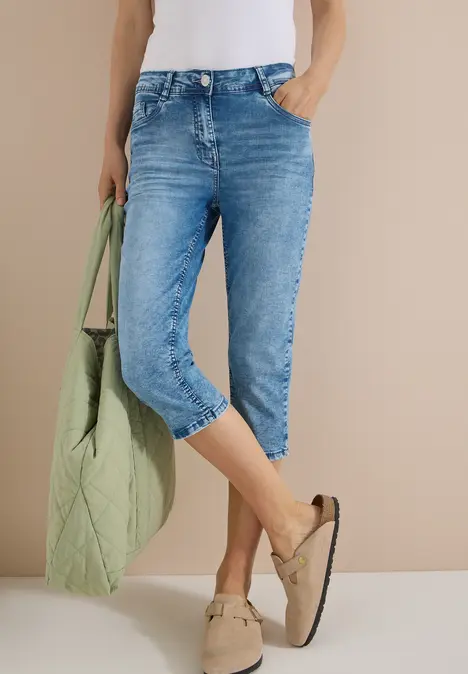 Eine Person trägt hellblaue, knielange Jeans, einen weißen Top, eine grüne Tasche und beige Mokassins.