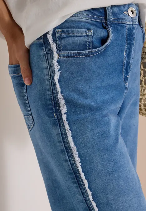 Blaue Jeans mit fransiger Seitennaht, Person trägt weißen Oberteil, Hand in Tasche.