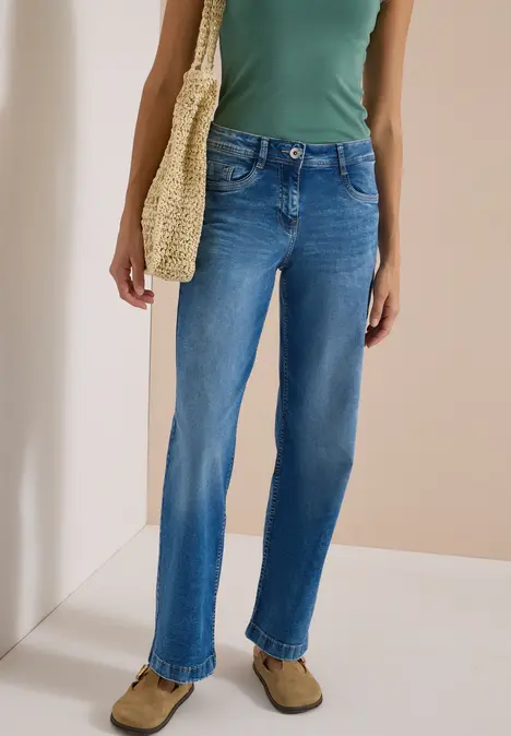 Eine Person trägt ein grünes Top, blaue Jeans und hält eine geflochtene Tasche.