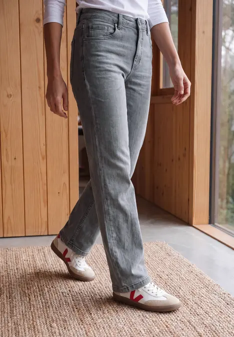 Beine in hellgrauen Jeans und weißen Sneakern mit rotem V-Logo, stehen auf Teppich am Fenster.