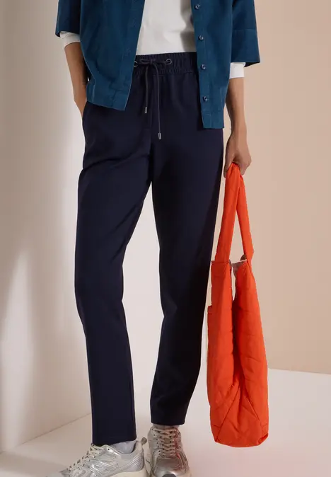 Person trägt dunkelblaue Freizeithose, weiße Sneaker und hält eine orangefarbene Tasche.