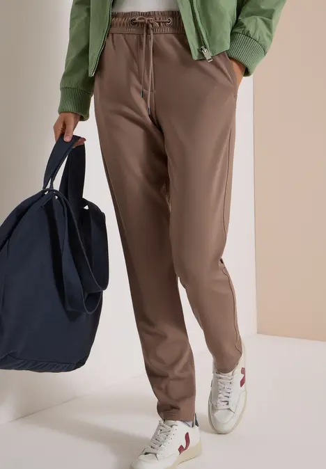 Modell trägt braune Jogginghose, weiße Sneaker mit Streifen, grüne Jacke und dunkelblaue Tasche.