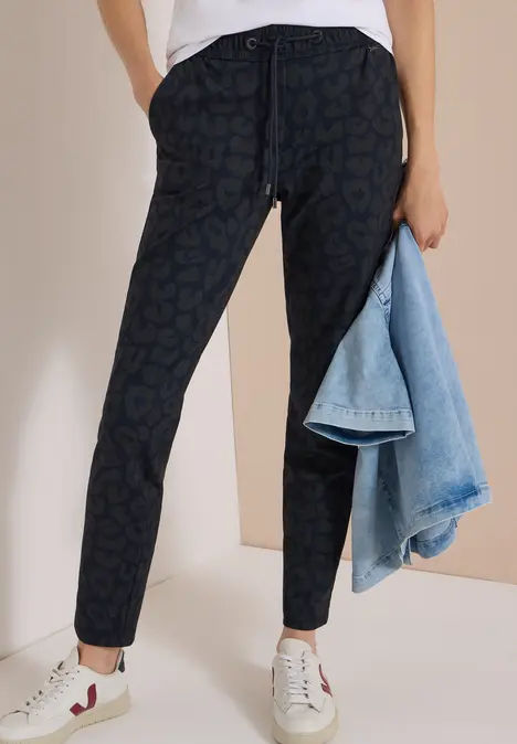 Dunkle Jogginghose mit dezentem Print, weiße Sneaker und hellblaue Jeansjacke als Accessoire.