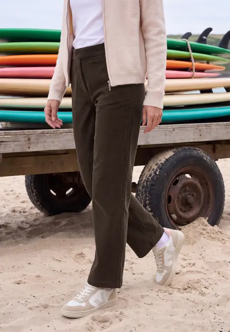 Eine Person trägt braune Cordhose, beige Jacke und weiße Sneaker an einem Sandstrand vor bunten Surfbrettern.