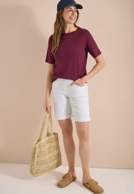 Frau trägt lila T-Shirt, weiße Shorts, Strohtasche und Loafer vor beigem Hintergrund.