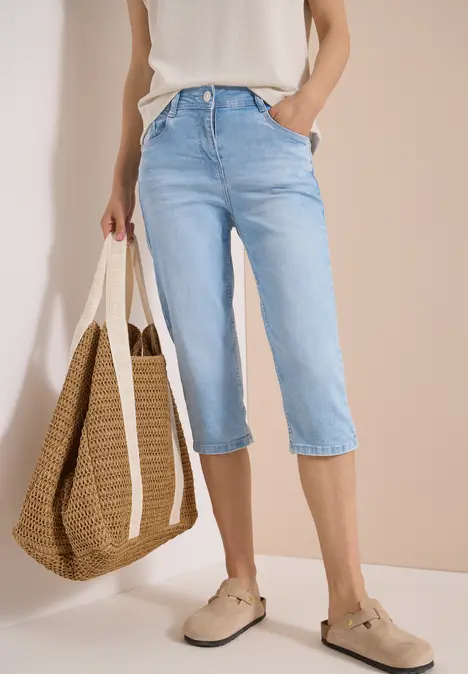 Frau in hellblauer Capri-Jeans mit weißem Oberteil, Basttasche und beigen Espadrilles vor neutralem Hintergrund.
