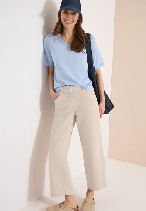 Eine Frau steht lässig an einer hellen Wand, trägt hellblaues T-Shirt, beigen Culotte-Hose, Cap und Umhängetasche.