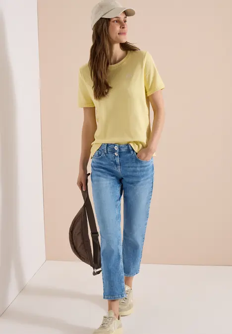 Frau in gelbem T-Shirt, blauen Jeans und Sneakern mit Cap, trägt Tasche locker an der Seite.