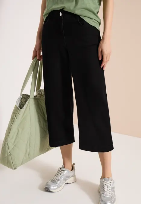 Eine Person trägt schwarze, weite Capri-Hosen, silberne Sneaker und hält eine grüne Stofftasche.