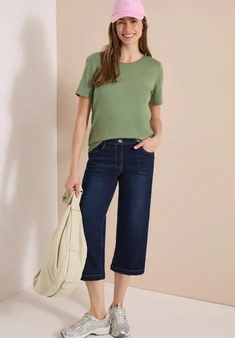 Person mit grünem T-Shirt, dunkelblauen Culotte-Jeans, weißen Sneakern und heller Tasche vor beigem Hintergrund.