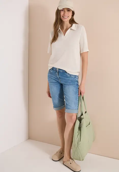 Frau in hellem Top, Jeans-Bermuda und großer grüner Tasche vor beiger Wand.