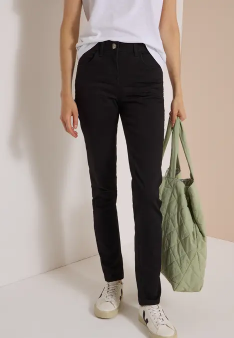 Person trägt schwarze Slim-Fit-Hose, weißes T-Shirt, weiße Sneaker und hält eine grüne gesteppte Tasche.