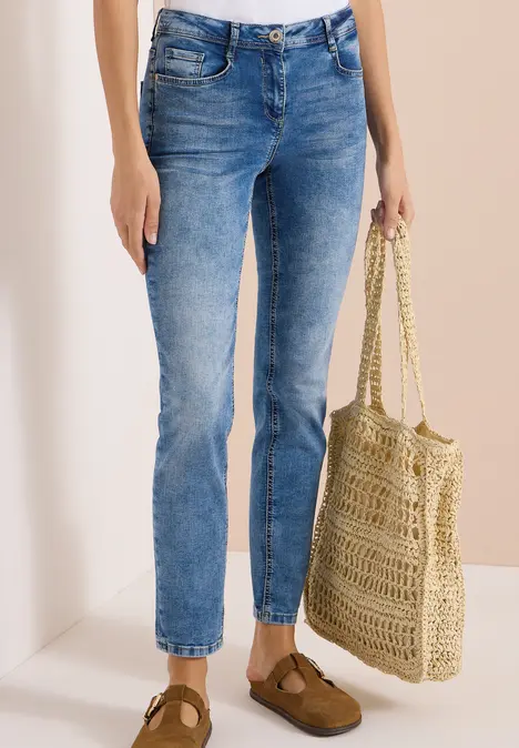 Frau trägt blaue Jeans, braune Schuhe und hält eine geflochtene Basttasche.
