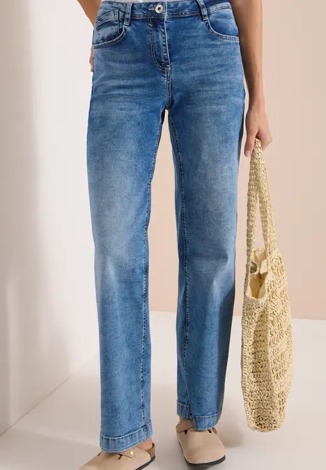 Person trägt hochgeschnittene, gerade geschnittene blaue Jeans, beige Ballerinas und hält eine Strohtasche.