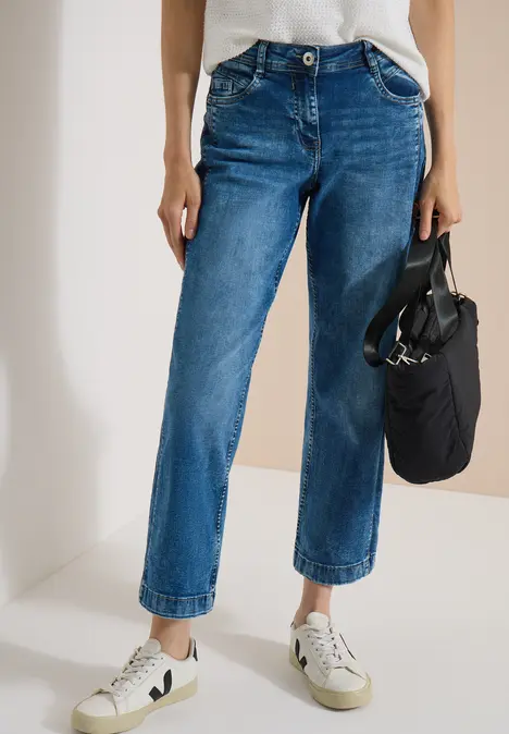 Frau trägt blaue, gerade geschnittene, hochgekrempelte Jeans mit weißen Sneakern und hält eine kleine schwarze Tasche.