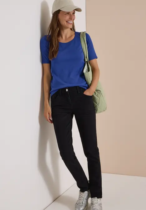Eine Frau lehnt lächelnd an einer Wand, trägt ein helles Cap, blaues Shirt, schwarze Hose und grüne Tasche.