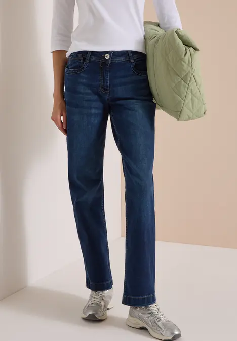 Person trägt dunkle Jeans, weißes Oberteil, silberne Sneakers und hält eine grüne gesteppte Tasche.