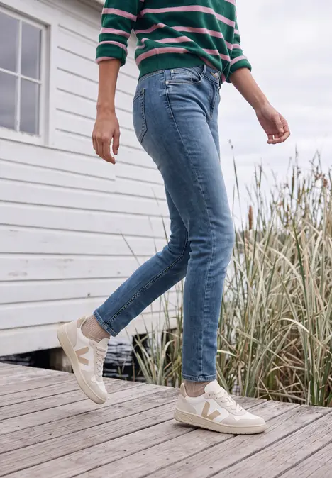 Eine Person geht auf einem Bohlenweg neben Dünen mit Gräsern, trägt blaue Jeans, gestreiftes Oberteil und weiße Sneaker.