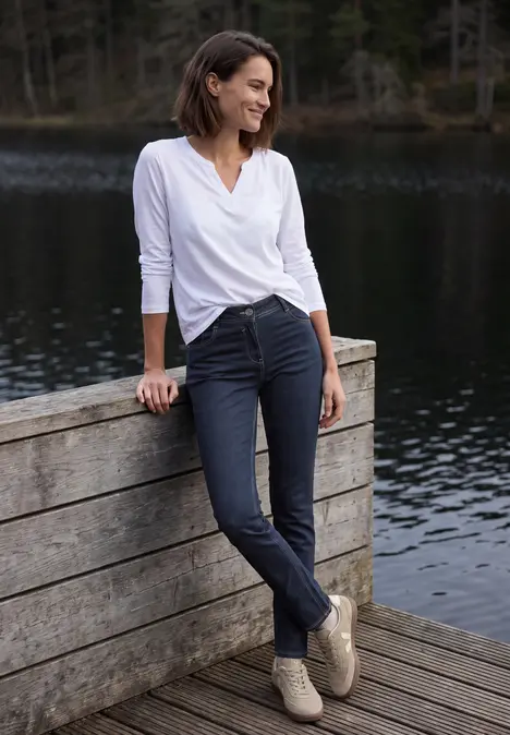 Eine Frau lehnt entspannt an einem Holzgeländer am See, trägt weiße Bluse, dunkle Hose und lächelt.