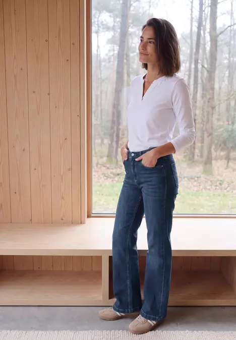Frau im weißen Hemd und weiten Jeans steht seitlich in einem hellen Holzraum vor einem Fenster mit Waldblick.