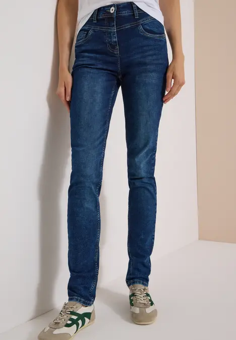 Person trägt schmale dunkelblaue Slim-Fit-Jeans und weiße Sneaker mit grünen Details vor schlichtem Hintergrund.