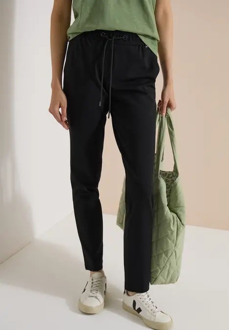 Dame trägt schwarze Jogginghose, weiße Sneaker, grünes Oberteil und gesteppte grüne Tasche.