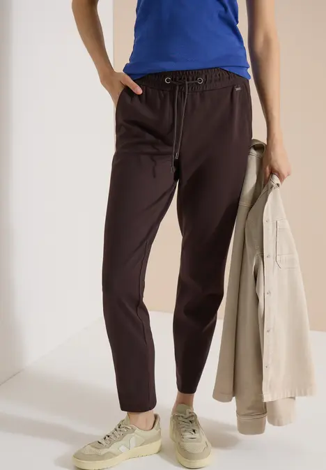 Frau trägt blaue Bluse, dunkelbraune Hose und helle Sneaker, hält eine beige Jacke in der Hand.