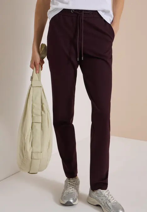 Person trägt eine weiße Bluse, bordeauxrote Hose, graue Sneaker und hält eine hellbeige Umhängetasche.