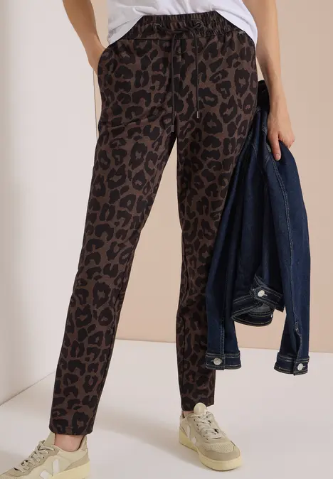 Frau in locker sitzender, braun-schwarzer Hose mit Leopardenmuster und Sneakern, hält Jeansjacke in der Hand.