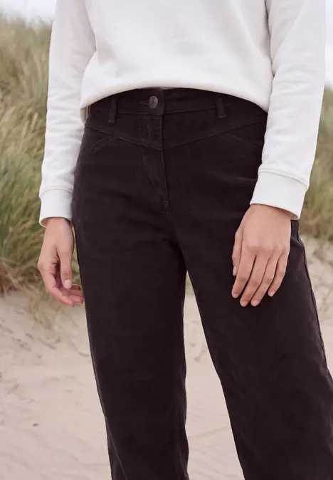 Eine Person steht im Sand, trägt weißen Pullover und schwarze Hose, im Hintergrund Dünen.