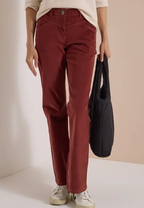 Eine Person trägt bordeauxrote Cordhose, weiße Sneaker und hält eine schwarze Tasche.