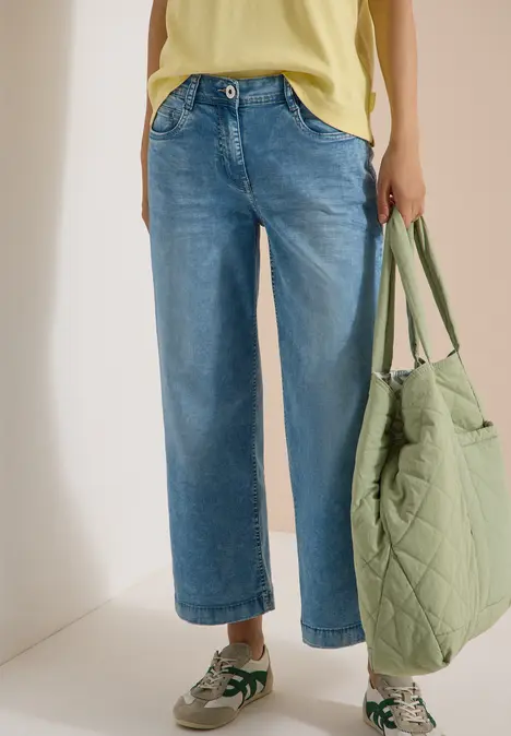 Person trägt weite blaue Jeans, gelbes Top, grüne Stepp-Tasche und Sneaker.