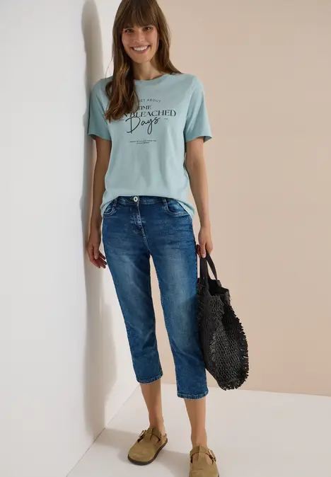 Frau in hellblauem T-Shirt, lässigen Jeans, grünen Slippern und schwarzer Korbtasche vor zweifarbigem Hintergrund.