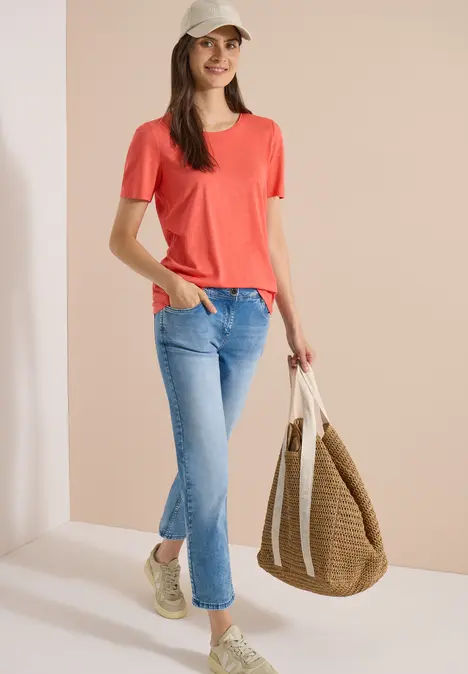 Frau trägt rotes T-Shirt, blaue Jeans, weiße Sneaker und hält eine geflochtene Tasche.