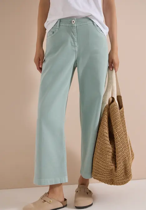 Person trägt weite, hellgrüne Hose mit hoher Taille, weißes Oberteil, bequeme Sandalen und hält eine geflochtene Tasche.