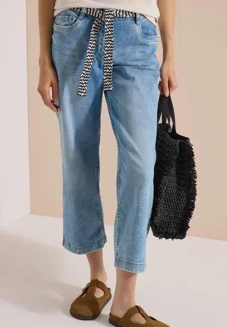 Frau in hellblauen Cropped-Jeans mit schwarz-weißem Gürtel, braunen Wildleder-Slippern und schwarzer Tasche.