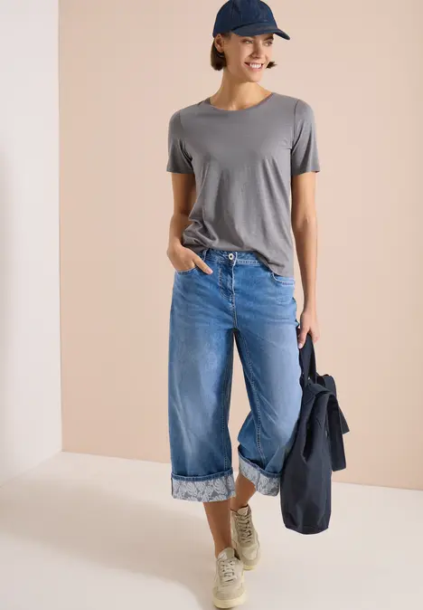 Frau in grauem T-Shirt, hochgekrempelte blaue Hose, Sneaker und Cap, hält eine Jeansjacke vor hellem Hintergrund.